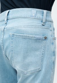 Κοντινό πλάνο πίσω τσέπης τζιν από ανοιχτό μπλε ύφασμα με ετικέτα επωνυμίας και ασημί καρφιά, φορεμένα με μαύρο τοπ.