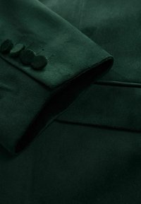 Ciemnozielona materiałowa rękawica o gładkiej fakturze, z pięcioma okrągłymi, teksturowanymi guzikami i starannie złożonym mankietem.