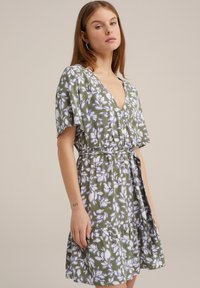 Rochie florală în nuanțe de verde cu modele albe și purpurii, cu mâneci scurte evazate, decolteu în forma de V și talie ajustată cu un detaliu de legare.