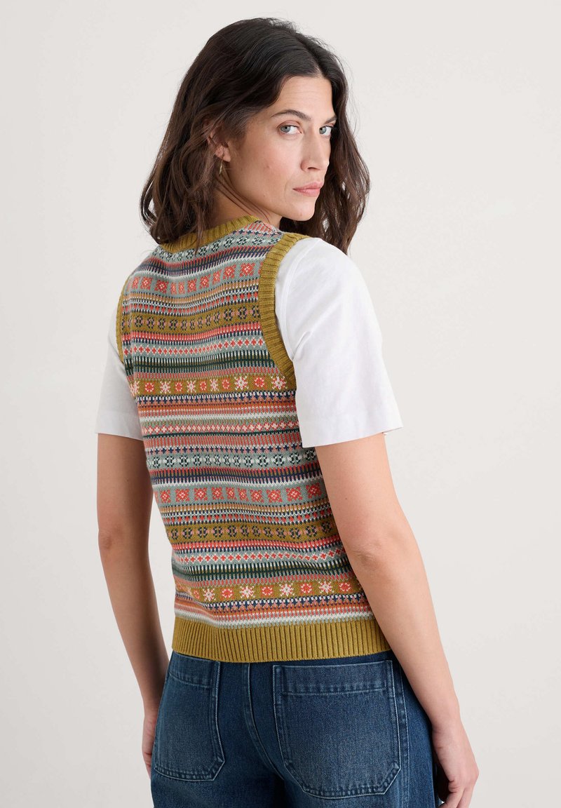 Gebreide vest met een patroon van meerkleurige geometrische ontwerpen, olijfgroene afwerking en een getailleerde pasvorm, gedragen over een wit T-shirt en blauwe jeans.