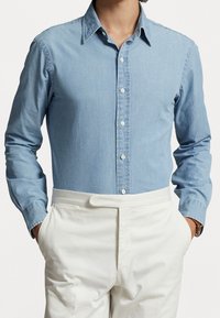 Ljusblå, långärmad denimskjorta med krage med knappar, knappslå fram och matchande knappar. Kombineras med vita byxor.