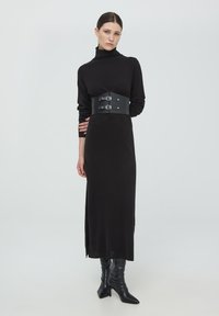 Abito lungo nero con maniche lunghe e colletto alto, vita sfiancata con cinture in finta pelle nera con accessori in argento. Stivali neri alti fino al ginocchio.