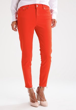 Schmal geschnittene, leuchtend rote Hose getragen mit blassrosa, hochhackigen Schnürschuhen und einer weißen Langarmbluse, gezeigt von der Taille bis zu den Füßen.