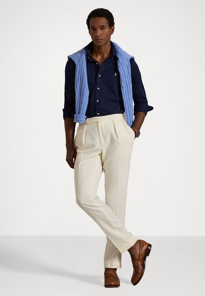 Ναυτικό πουκάμισο, ανοιχτό μπλε πλεκτό γιλέκο, κρέμα πλισέ παντελόνια και καφέ δερμάτινα παπούτσια, τονίζοντας μια κλασική, ραμμένη εμφάνιση.