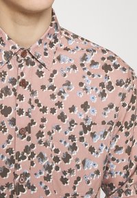 Gros plan d'une personne portant une chemise rose à boutons avec un motif floral marron et bleu et une poche poitrine frontale.