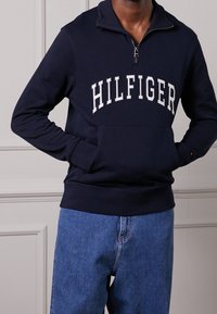 Homme portant un sweat à col zippé marine avec le texte "HILFIGER" et un jean bleu, les mains dans les poches du sweat, debout à l'intérieur.