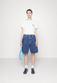 Hvit kortermet t-skjorte med blå delfin grafikk, kombinert med kne-lange blå denimshorts. Holder en turkise strikket veske, iført marineblå joggesko.