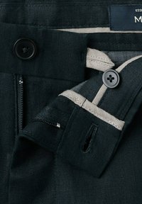 Nærbillede af taljebånd på sorte bukser med knap, lynlås og indvendig kontrasterende beige stofdetalje.