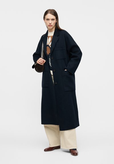 Γυναίκα που φοράει ένα μακρύ σκούρο ναυτικό παλτό πάνω από ένα φλοράλ πουλόβερ, φαρδιά μπεζ παντελόνια, καφέ παπούτσια και κρατά μια μικρή σκούρα χειραποσκευή στον ώμο.