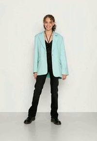 Blazer oversized azul claro, blusa preta, calças pretas, com uma textura suave. Modelo posa confiante com as mãos nos bolsos num fundo neutro.