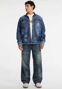 Veste en denim avec deux poches poitrine et teinte bleue délavée, associée à un jean cargo avec plusieurs poches. Des baskets décontractées complètent la tenue.