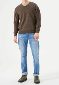 Pull marron en V avec un tricot texturé ; associé à un jean bleu clair, retroussé aux chevilles, et des baskets vertes. Logo sur la poitrine du pull.