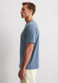 Helles blaues T-Shirt mit kurzen Ärmeln und Rundhalsausschnitt, aus einem weichen Stoff gefertigt, mit einer subtilen Textur und einem Logodetail am Saum.
