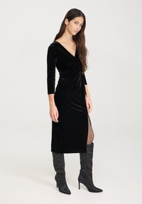 Czarna aksamitna sukienka z rękawami o długości trzy czwarte, dekoltem w serek, skręconym detalem z przodu i rozcięciem po bokach, zestawiona z wysokimi na kolana czarnymi botkami.