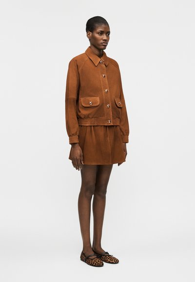 Γυναίκα που στέκεται φορώντας καφέ σακάκι με κουμπιά και τσέπες με καπάκια, ταιριαστό φούστα και φλατ παπούτσια με λεοπάρ μοτίβο σε απλό φόντο.