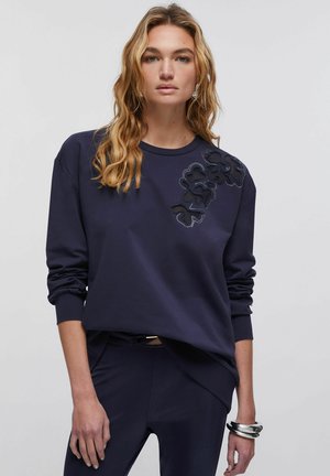 Donna con capelli biondi ondulati indossa una felpa blu navy con ritagli floreali texturizzati sulla spalla e pantaloni blu navy abbinati, posando con un braccialetto d’argento.