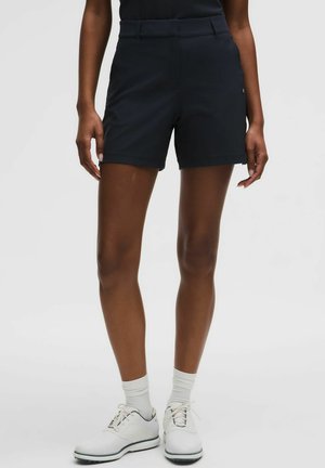 Persona que lleva pantalones cortos negros a medida, calcetines blancos y zapatos blancos con cordones, de pie contra un fondo claro y liso.