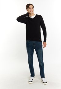 Pulover negru din tricot cu strițe, cu decolteu în V, asortat cu blugi albaștri și adidași albi. Bărbatul se pozează cu o mână pe gât, zâmbind.