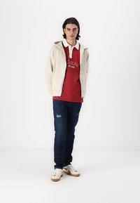 Red polo shirt with white collar, beige hoodie, blue denim jeans with distressed details, and white sneakers with brown accents.
