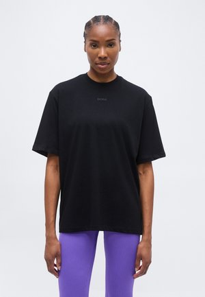 Kobieta stojąca, ubrana w luźną czarną koszulkę z napisem "BORG" oraz jaskrawo fioletowe prążkowane legginsy, na tle białej ściany.