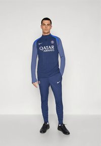 Tuta da allenamento blu navy con texture a coste, colletto alto, accenti blu e logo Qatar Airways. Scarpe sportive nere completano l'insieme.