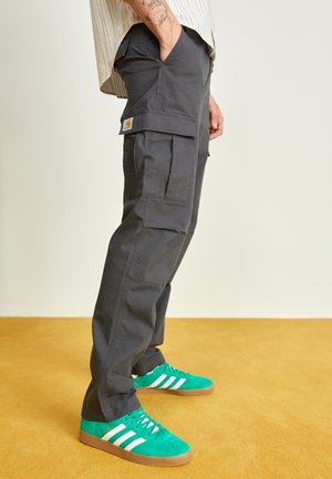 Personne portant un pantalon cargo gris foncé et des baskets Adidas vertes avec des rayures blanches, debout sur un sol jaune moutarde.