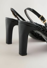 Sandales à talons hauts noires avec des brides tressées et des boucles dorées réglables sur un fond clair, inclinées pour montrer les talons et les brides.