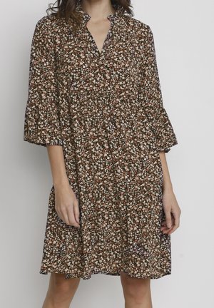 Frau trägt ein knieumspieltes, locker sitzendes Kleid mit dreiviertellangen Ärmeln und einem dunklen Blumenmuster in Weiß-, Braun- und Rosatönen.