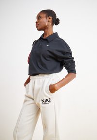 Fekete, hosszú ujjú poloing, rajta egy kis Nike logóval, világos bézs sportnadrággal kombinálva, amelyen szintén van Nike logó. A modell egyik kezét a zsebébe dugva áll.