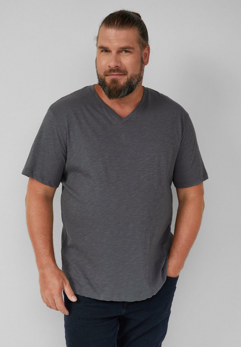 Szara krótka koszulka z dekoltem w serek, wykonana z lekkiego, teksturowanego materiału. Posiada luźny krój i subtelny melanżowy wzór. Noszona z ciemnymi dżinsami.