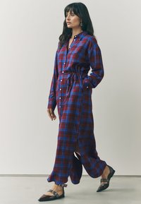 Rød og blå ternet lang skjorte-kjole med bælte, knaplukning foran, lange ærmer, sideslids, brugt med sorte spidse ballerinaer. Glat stofstruktur.