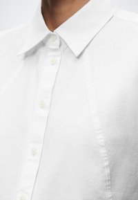 Κοντινό πλάνο ενός ατόμου που φοράει ένα λευκό πουκάμισο με κουμπιά και μυτερό γιακά, εμφανίζοντας το άνω μέρος του στήθους και τον λαιμό.