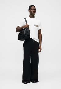 Hvit t-skjorte med korte ermer og trykt lommendesign, kombinert med løse svarte bukser og en svart veske med teksturerte detaljer.