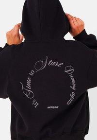 Zwarte hoodie van zachte stof met een grote, gebogen witte tekstprint op de achterkant. Bevat een klein logo aan de onderzijde.