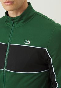 Groene zip-up jacket met zwart paneel en witte bies. Heeft een gestructureerde stof en een klein geborduurd logo op de borst.