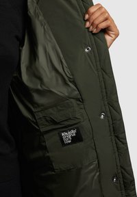 Jachetă matlasată de culoare verde măslinie, cu interior și patch negru. Prezintă închideri cu capse și un buzunar lateral. Textură netedă și lucioasă.