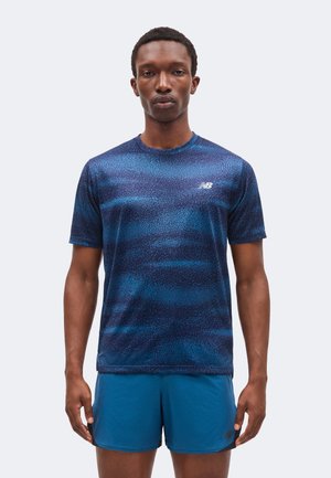 Uomo con maglietta sportiva blu a maniche corte fantasia e pantaloncini blu tinta unita, in piedi davanti a uno sfondo chiaro uniforme.