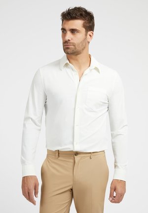 Camicia bianca a maniche lunghe con colletto classico, taschino singolo sul petto e chiusura con bottoni, abbinata a pantaloni color cammello. Tessuto morbido.