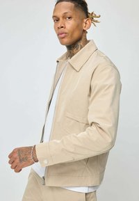 Jeune homme tatoué portant une veste beige fermée sur une chemise blanche, se tenant devant un fond léger uni.