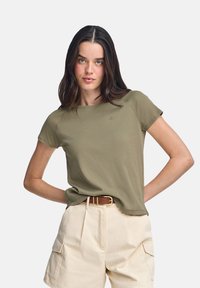 T-shirt verde oliva a maniche corte con scollo rotondo e un discreto dettaglio del logo sul petto, abbinato a pantaloni cargo beige chiaro.