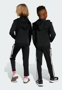 Två barn står med ryggen mot kameran iförda svarta huva-jackor och svarta träningbyxor med vita ränder, tillsammans med Adidas-skor.