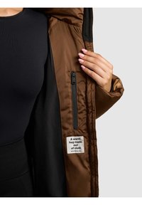 Personne tenant ouverte une veste marron montrant une poche intérieure zippée et une étiquette portant l'inscription « Un câlin chaleureux fait de tissu. »