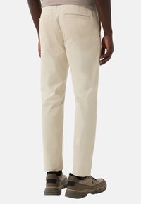 Homme portant un pantalon beige coupe droite et des baskets marron, vu de dos contre un fond blanc uni.