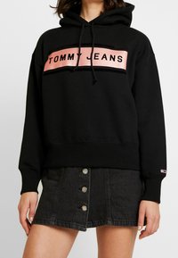 Mujer vestida con una sudadera con capucha negra que tiene el logo "TOMMY JEANS" en rosa y una falda de mezclilla negra con botones al frente, de pie contra un fondo blanco.
