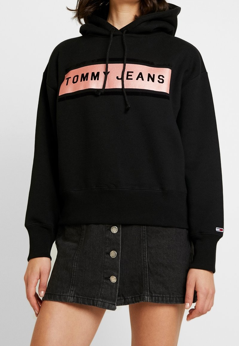 Mujer vestida con una sudadera con capucha negra que tiene el logo "TOMMY JEANS" en rosa y una falda de mezclilla negra con botones al frente, de pie contra un fondo blanco.