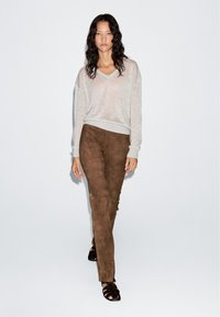 Woman with long curly hair walking, wearing light beige sweater, brown suede pants, and black open-toe sandals against a white background.