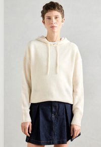 Κρέμα φωτεινής απόχρωσης πλεγμένη κουκούλα με χαλαρή εφαρμογή, διαθέτοντας κορδόνι στην κουκούλα και λάστιχο στις μανσέτες, σε συνδυασμό με μια σκούρα τζιν φούστα.