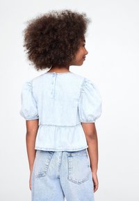 Lys blå denimtopp med pufferm, innsvingt midje og knappelukking bak; brukt sammen med lys blå denimjeans.