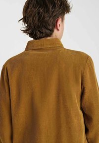 Persoon met kort bruin haar, gekleed in een mosterdgele corduroyjas, gezien van achteren tegen een effen lichte achtergrond.