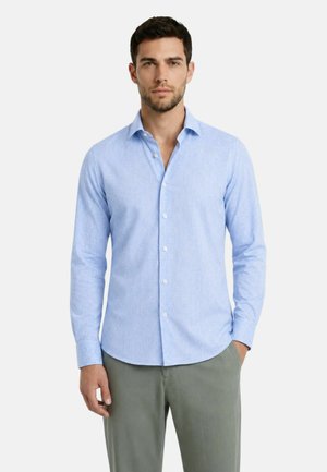 Uomo che indossa una camicia azzurro chiaro con bottoni e pantaloni verde oliva, in piedi con un’espressione neutra e una mano in tasca.